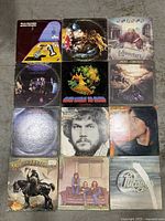 Top-down view of 12 vinyl LP album covers arranged in 3 rows by 4 columns on a concrete floor, showing worn and rough jackets.
