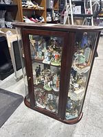 Full view of cherry wood and glass wall cabinet filled with colorful Norman Rockwell figurines on three shelves.