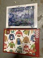 Two unopened 1000-piece boxes of jigsaw puzzles, one featuring Claude Monet's artwork and the other with Christmas sweaters design.