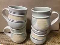 Four ceramic mugs stacked in pairs, showing two designs: one with navy-blue horizontal stripes, the other with a ribbed texture and light-colored leaf motif.