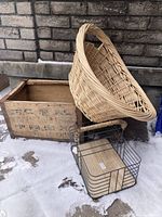 Full lot showing vintage wooden crate with Chinese characters, a large oval wicker basket resting on crate, and wire carry basket in front on snow-covered ground.