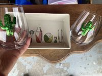 Close-up of two stemless glasses with green cactus decoration and wooden tray with a white ceramic inset with cocktail illustrations.