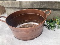Oval copper-like bucket with two handles placed on snow, showcasing shape and color.