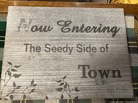 Photo of a rectangular wooden garden sign with tin lettering saying 'Now Entering The Seedy Side of Town'. Botanical silhouettes are visible on the bottom corners.