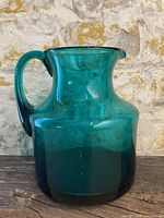 Photo showing the full blue glass mini pitcher with handle and spout against a stone wall background.