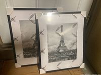 Two new black border IKEA picture frames with Eiffel Tower prints, in packaging with protective corners, placed upright on the floor.