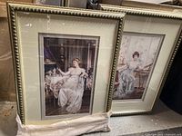 Two framed art prints featuring vintage-style ladies in white dresses, displayed side by side on the floor, leaning against a wall.