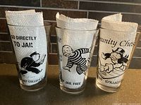Three Monopoly-themed drinking glasses with visible printed characters and slogans placed side by side.