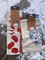 Three new cotton produce bags with colorful prints and tags laid on snow, drawstring visible