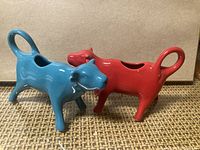 Red and blue ceramic cow-shaped creamers with tail handles and top openings for cream pouring.