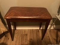 Photo showing a wooden piano bench with brown cushioned seat against a beige wall and wooden floor