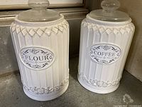 Two off-white ceramic canisters side by side labeled FLOUR and COFFEE with glass lids on a countertop.