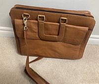 Front and side view of brown leather briefcase-style bag standing on carpeted floor showing shape, handles, and front pocket.