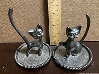 Two silver tone cat figurine ring holders on a dark wooden surface with a ruler behind to show height approx 4 inches