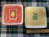 Photo showing two square ceramic fruit plates with colorful backgrounds and labeled fruit illustrations.