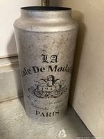 Tin vase with Paris Cafe De Madaline 1932 design. Shows front and details of printed text and graphics on metallic silver finish.