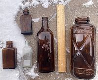 Five vintage glass bottles on a snowy concrete surface next to wooden ruler showing scale. Includes one large amber jar with embossing, one amber bottle with embossed figure, two smaller amber bottles, and one clear bottle.