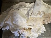 Photo showing assorted white and cream vintage lace linens including tablecloths and doilies, showcasing scalloped edges and intricate lace patterns.