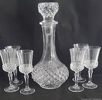 Full lot view showing crystal decanter with stopper surrounded by six matching stemmed sherry glasses on black background.