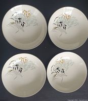 Top view of four ceramic coupe bowls arranged in square, showing decorative botanical leaf patterns in gold, black, and gray on cream background.