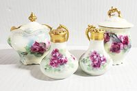 Set of six vintage porcelain condiment items; includes salt and pepper shakers, two condiment pots with lids, and two spoons, all decorated with pink and purple floral hand painting and gold accents on lids and handles.