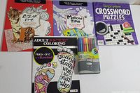 Four adult activity books and a pack of 18 felt tip markers arranged on a surface, showing front covers and packaging of markers.