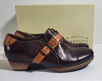 Side view of the left shoe showing dark brown leather upper with light brown strap buckle detail and stacked heel against a neutral background with box