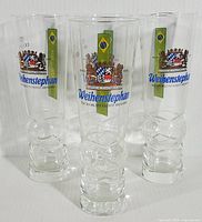 Four clear beer glasses standing upright showing front facing Weihenstephan logos with Brazil 2014 World Cup design and crest.