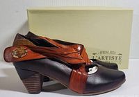 Side view of left shoe showing black leather body with brown leather ankle strap and wooden button detail. Stacked wooden heel shown with box in background.