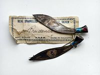Top view of two silver leaf-shaped brooches with original worn paper tag stating 'Real Enamelled Silver' and 'Made in Calcutta'. Brooches have colored enamel on narrow ends and engraved details.
