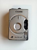 Front view of Canon ELPH LT camera showing silver faceplate with Canon lens and branding, visible wear and scratches