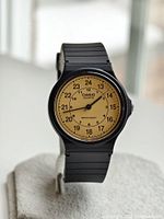 Front view of Casio MQ-24 wristwatch on display stand showing beige dial with 24-hour markers and black plastic strap.