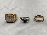 Lot of three sterling silver rings shown together on a neutral background. From left to right: a chunky signet ring with a textured surface, a delicate ring with a black heart stone and marcasite, and a patterned band ring.