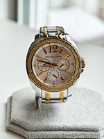 Front view of Michael Kors watch on stand showing silver and gold-tone metal with crystal bezel around the dial and three subdials on face.