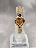 Full frontal view of the vintage Gruen ladies asymmetric wristwatch on clear display stand showing rhombus-shaped gold dial and gold-tone hands.