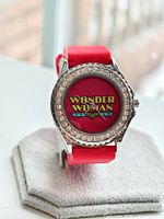 Front view of the Wonder Woman logo watch face with decorative bezel and red strap displayed on a watch cushion.