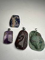 Four necklace pendants displayed flat on white surface: amethyst pendant in gold plated frame, horse carved stone pendant, wolf carved stone pendant, blue and white patterned rectangular stone pendant.