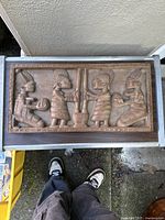 Top view of the full rectangular relief carving showing four figures in traditional dress engaged in pounding a mortar and holding gourds, edges framed with carved border.