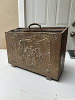 Front view of the brass hand tool box showing the detailed embossed scene of people in a village setting with trees and buildings.