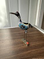 Full side view of the cloisonné bird incense burner standing on a wooden table with metal legs attached to a metal base with an orange sticker.