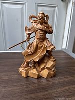 Front view of faux carved wood statue showing standing warrior figure detailed in armor and flowing cloth on rocky base