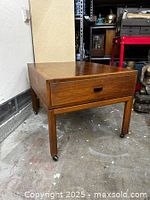 Full view of the nightstand showing its rectangular top, drawer with handle, four legs and caster wheels on a concrete floor.
