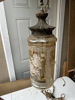 Side view showing cylinder-shaped painted glass with floral and border designs surrounding the lamp interior. Top metal piece in aged finish with chain for hanging.