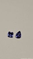 Photo showing two natural tanzanite gemstones side by side on a beige background, one pear-shaped and one cushion-shaped.
