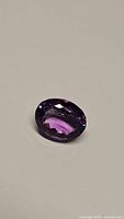 Close-up photo of deep purple, oval cut natural Brazilian amethyst gemstone with facets clearly visible.
