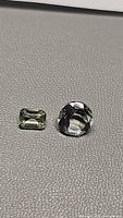 Two natural prasiolite gemstones on a textured grey surface, one emerald cut rectangular and one round diamond cut stone.