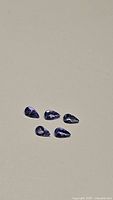 Five pear-shaped blue-purple Tanzanite gemstones arranged on a neutral background, showing size and shape.