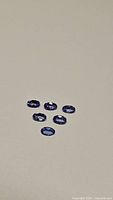 Six calibrated oval cut purple blue Tanzanite gemstones placed on plain light surface. Each stone approximately 5x3mm, showing purple blue coloration and high clarity consistent with VVS to flawless grade.