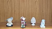 Four ceramic figurines displayed on a wooden surface against a wooden panel background: woman's head with hat, cow figurine, pine cone, and dog statue.