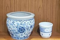 Photo showing both the large chinoiserie planter and the small blue and white floral planter side by side on a wooden floor with wood panel background.